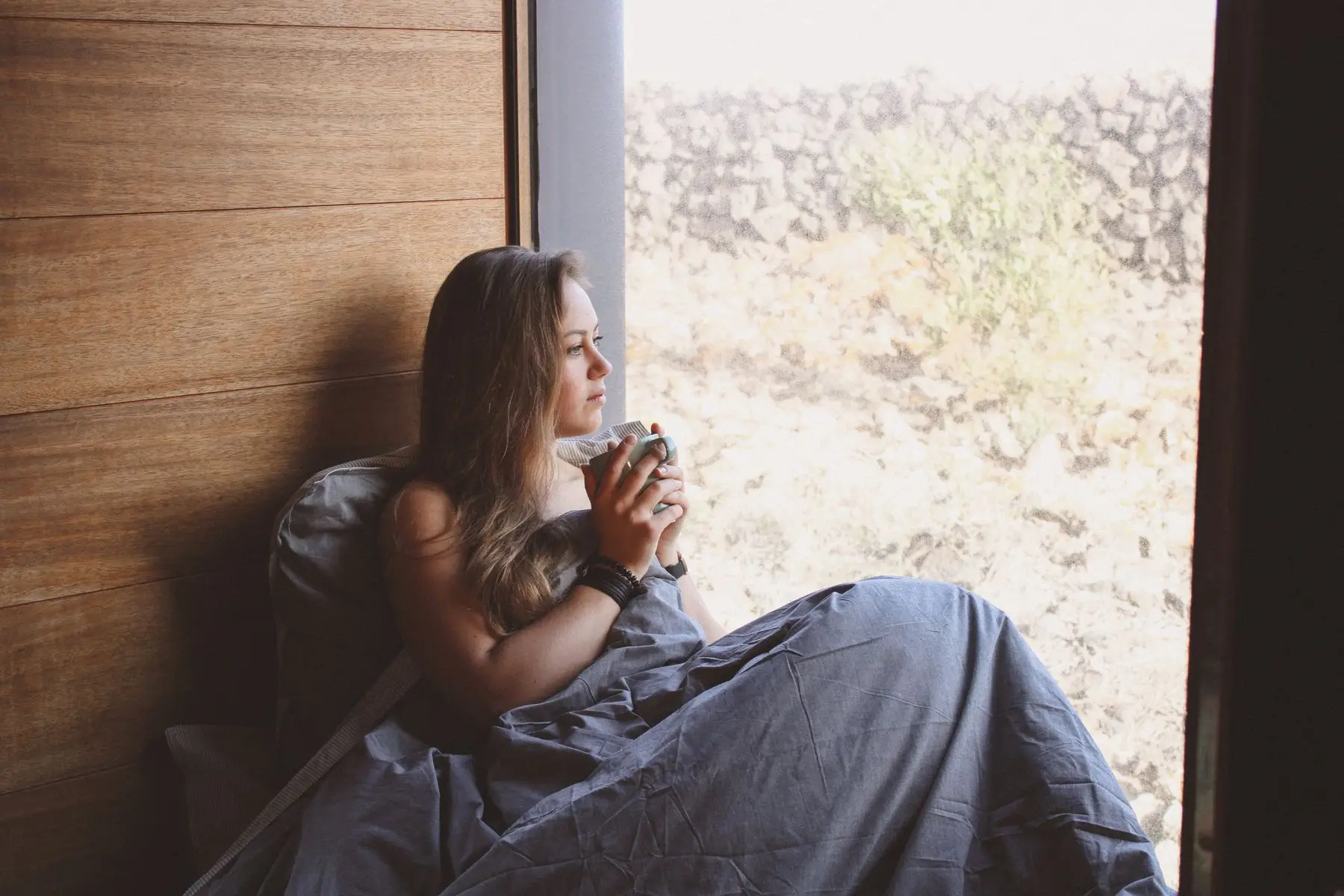 Woman wrapped in a blanket sitting by a window and holding a cup of tea in a quiet moment at home