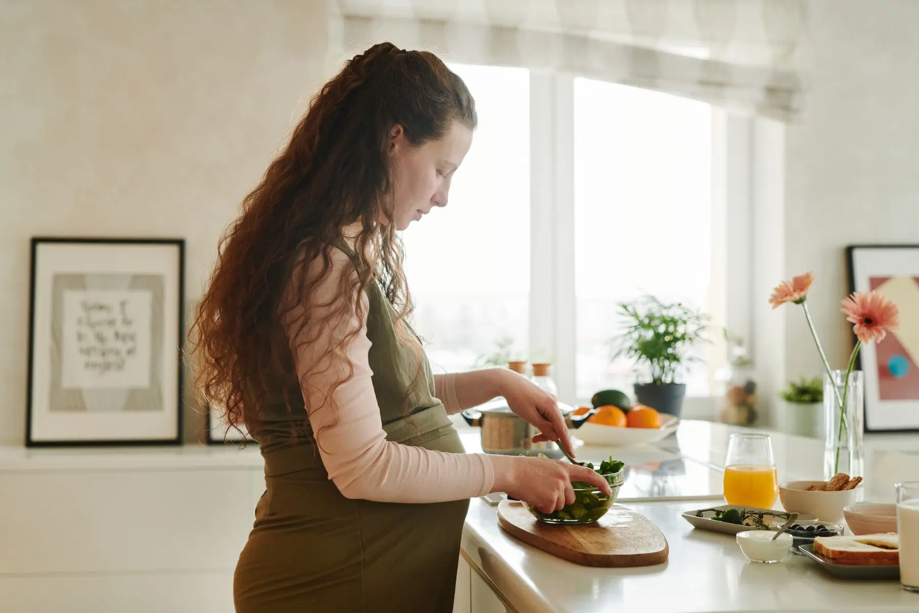 Pregnant woman with long curly hair preparing a fresh green salad at a bright kitchen counter