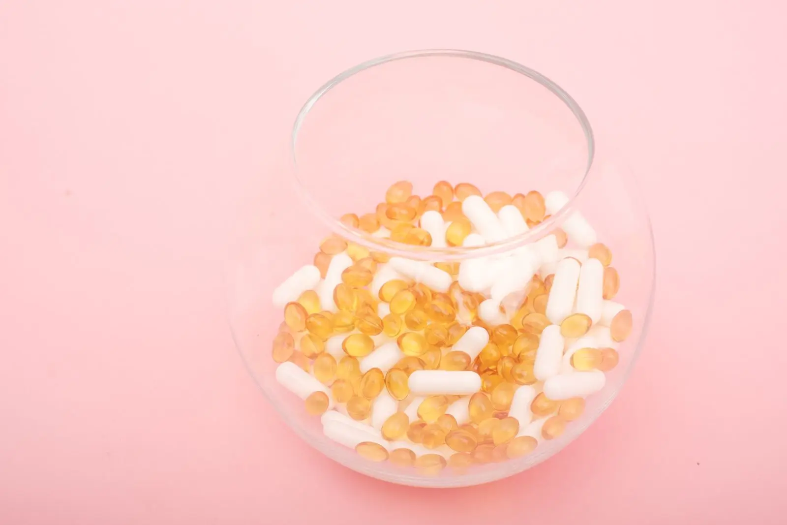 Assorted vitamin capsules in a glass bowl on a pink surface