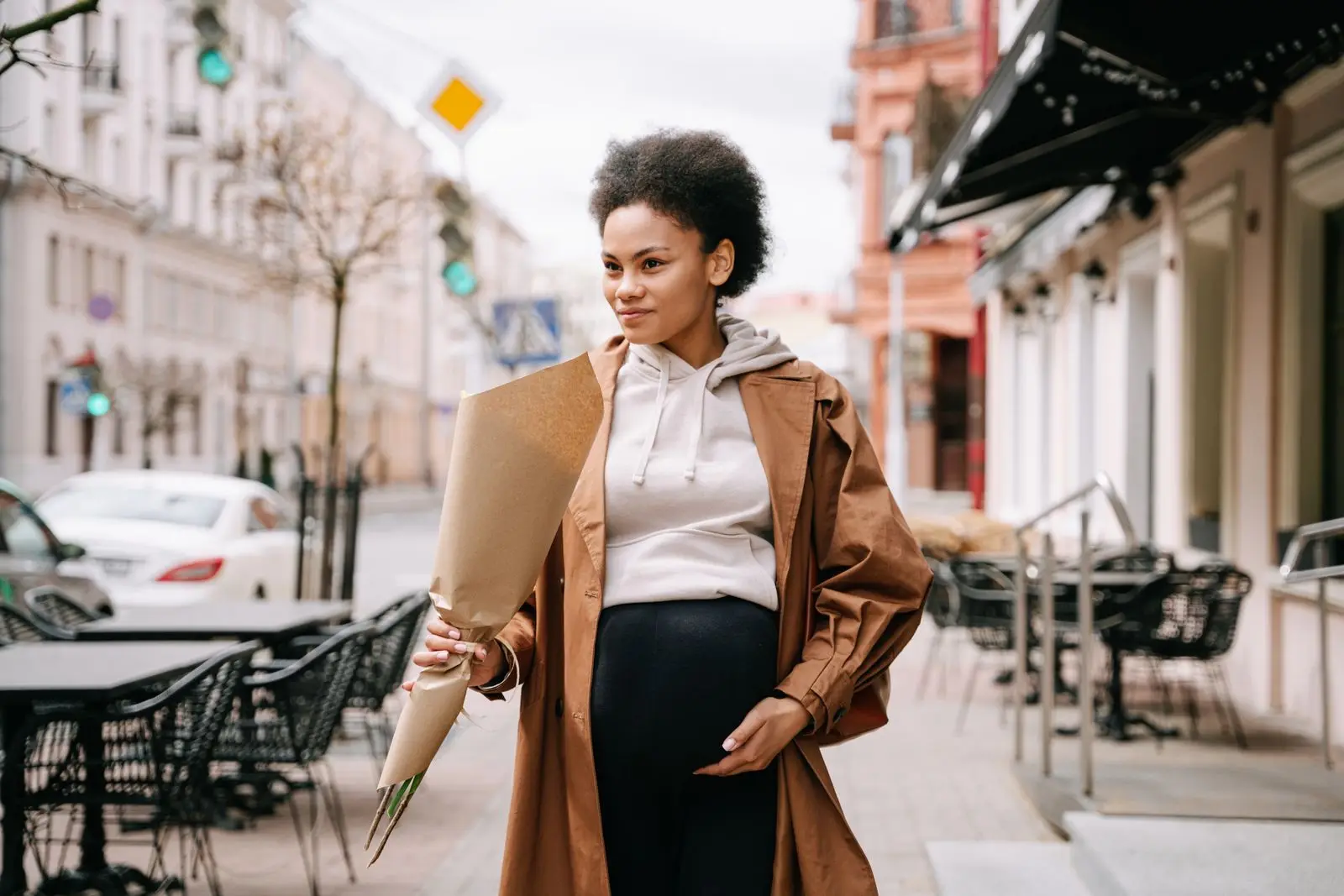 Pregnant woman in a brown sweater walking down a city street holding flowers